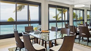 Un espace repas avec une table et des chaises en verre, de grandes fenêtres offrant une vue sur l’océan et un éclairage moderne suspendu au plafond.