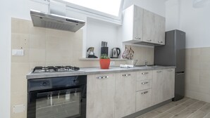 A modern kitchen with a gas stove, oven, and stainless steel refrigerator. It features light-colored cabinetry and a small window above the sink. - Appartamento Ecate (Ragusa)