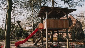 Een speeltuin met een houten glijbaan en klimconstructies, omringd door bomen en een bakstenen gebouw op de achtergrond.