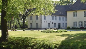 Een gebouw met een symmetrische gevel, meerdere ramen en een verzorgd gazon met enkele bomen en een bank.