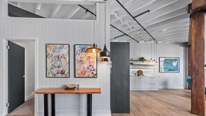 Un intérieur moderne avec un banc en bois, deux tableaux abstraits, un luminaire suspendu et un coin cuisine à l’arrière-plan.