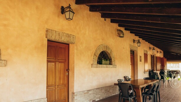 A covered outdoor dining area with a long wooden table and chairs, stone archway, and wooden doors.