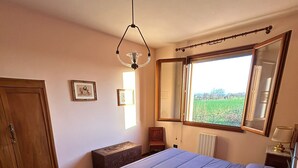 A bedroom with a wooden floor, a bed with a blue bedspread, a bedside table with a lamp, a wardrobe, and a window with a view of a field.