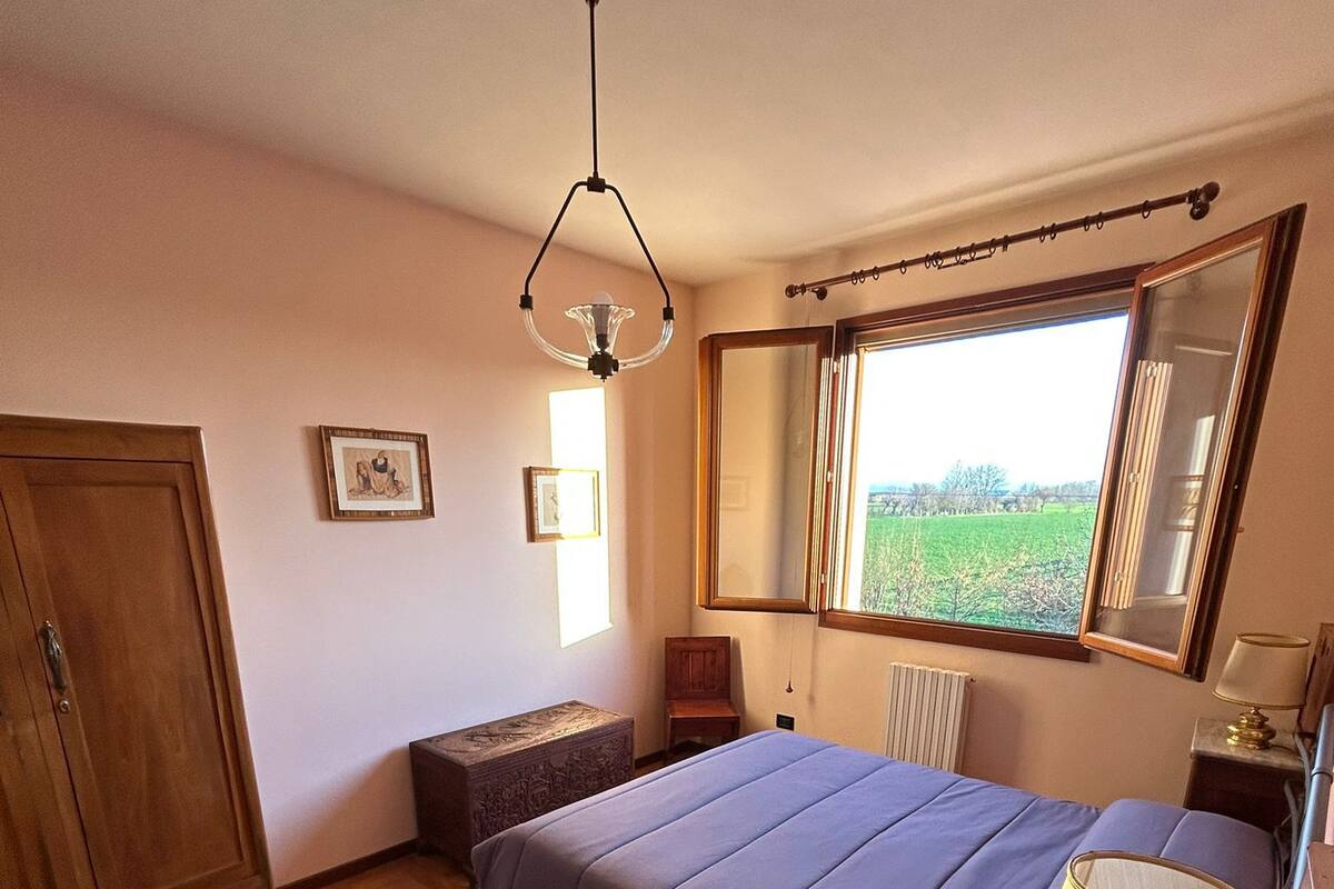 Ett sovrum med trägolv, en säng med ett blått överkast, ett sängbord med en lampa, en garderob och ett fönster med utsikt över ett fält.
