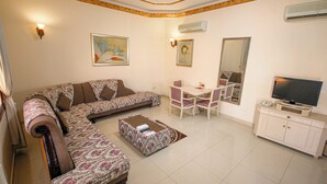 Un salon comprenant un canapé, un pouf, une table à manger avec des chaises, un téléviseur posé sur un meuble et des œuvres d’art accrochées aux murs.