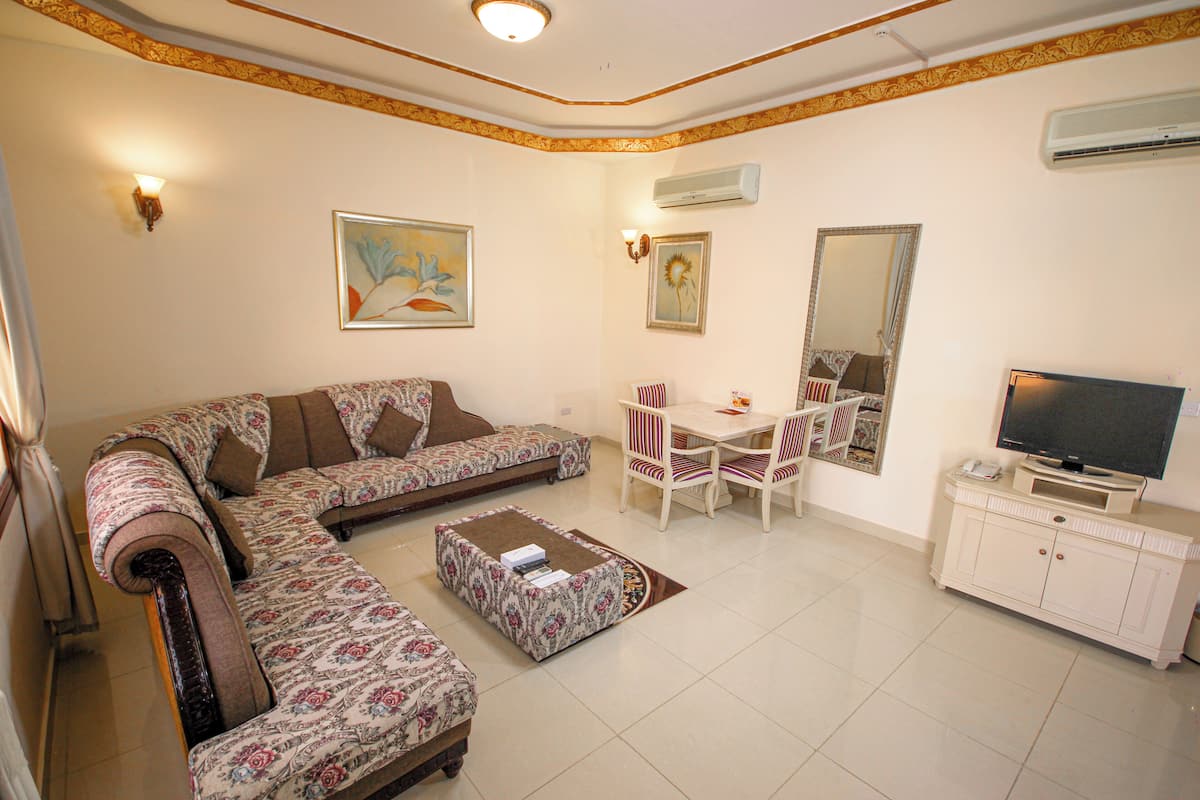 A living room with a sofa, ottoman, dining table with chairs, a TV on a cabinet, and artwork on the walls.