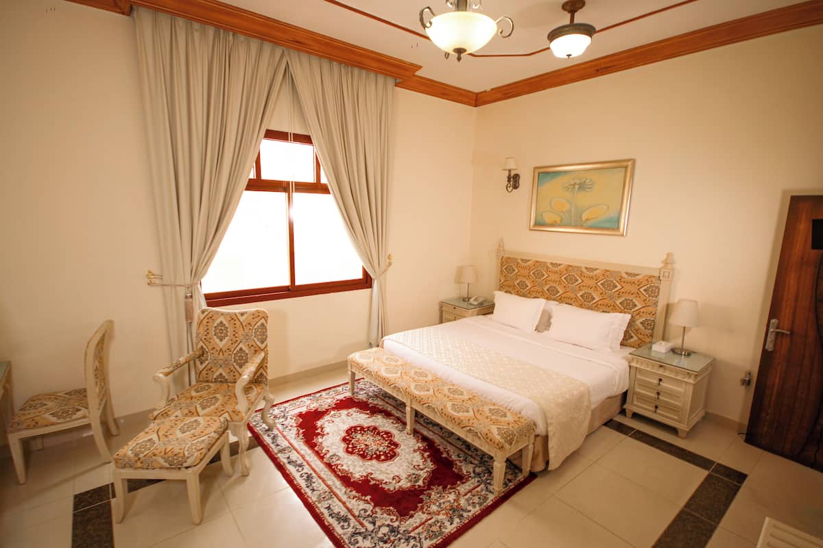 A bedroom with a large bed, a nightstand, a chair, a window with curtains, and a painting on the wall.