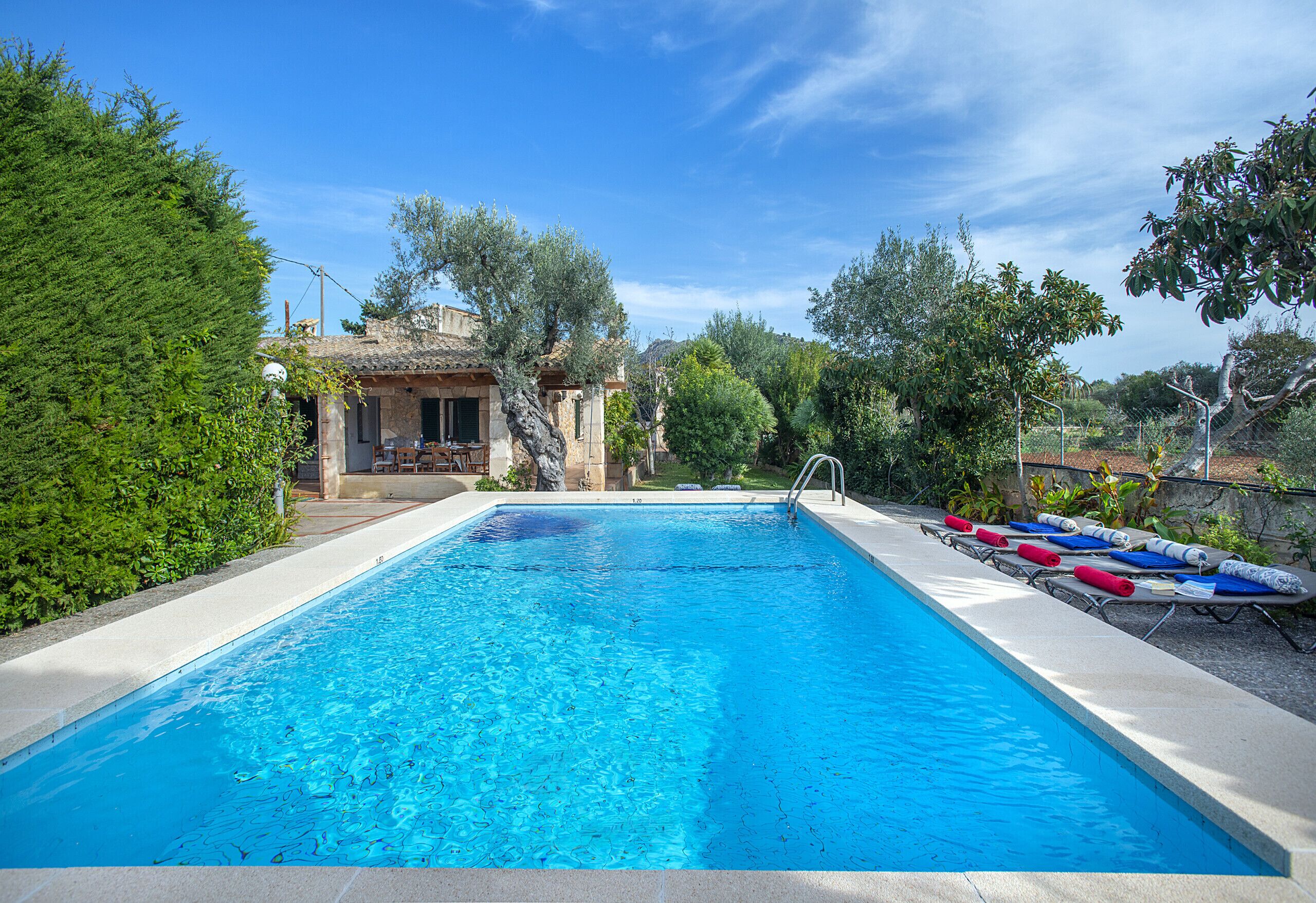 A rectangular swimming pool with a clear blue water surface, surrounded by a concrete deck. There are lounge chairs with towels on the right side of the pool.