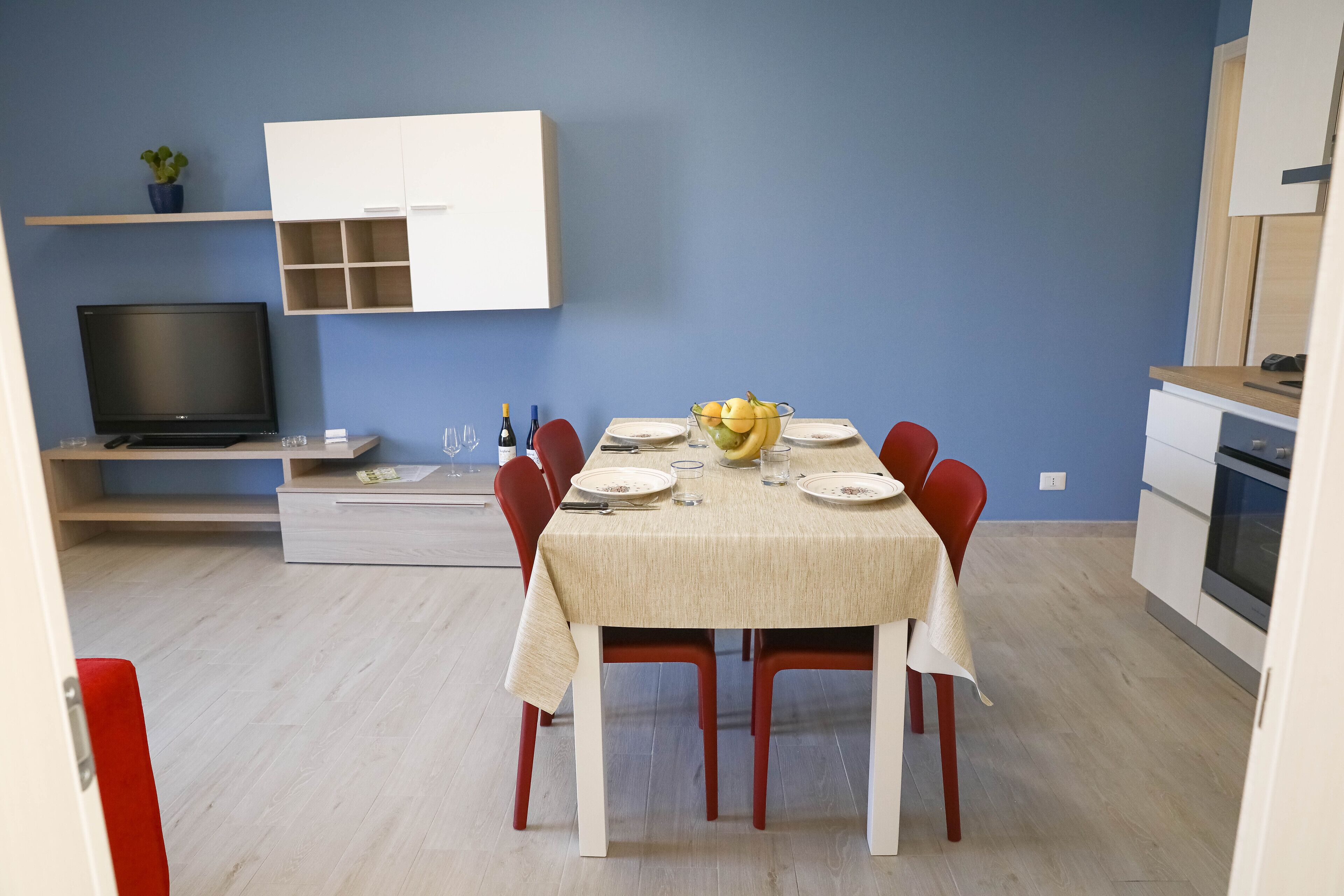 Een eetgedeelte met een tafel voor vier personen, een televisie, en een keuken met kastjes en een magnetron.