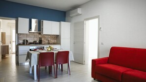A modern kitchen with white cabinets, a dining table with four chairs, and a red sofa in a living area. - Appartamento Orfeo (Ragusa)