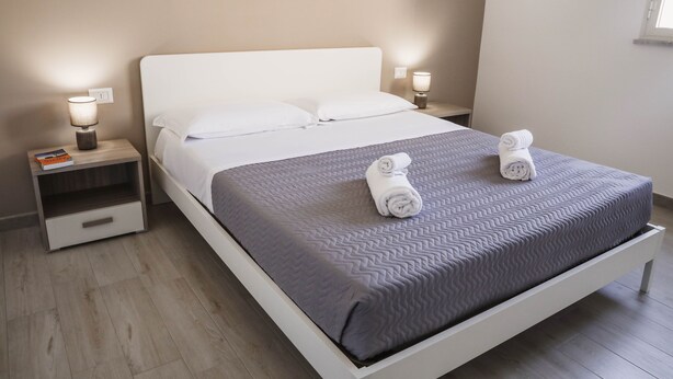 A neatly made bed with a grey quilt, two white pillows, and two rolled towels on the bed. There are two bedside tables with lamps, one on each side of the bed. - Appartamento Orfeo (Ragusa)