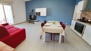 A modern living room with a red sofa, a dining table set for four, and a kitchen area with a stove and oven. - Appartamento Orfeo (Ragusa)