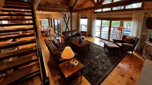 A spacious living room with a wooden ceiling, leather sofas, a coffee table, and a large window providing a view of the outdoors. - The Lake House on Otisco Lake, stunning family getaway (Marietta)