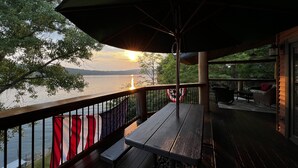 A wooden deck with a table and chairs, overlooking a body of water with a sunset in the background. - The Lake House on Otisco Lake, stunning family getaway (Marietta)