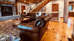 A spacious living room with a stone fireplace, leather sectional sofa, and a wooden staircase. - The Lake House on Otisco Lake, stunning family getaway (Marietta)