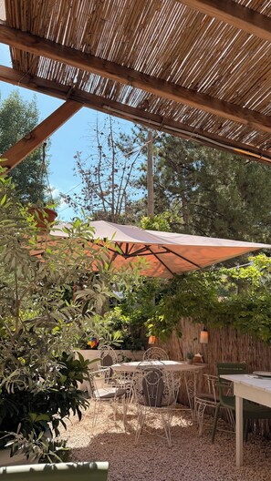 An outdoor patio with a thatched roof, a table and chairs, and surrounding greenery.