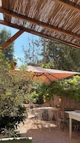An outdoor patio with a thatched roof, a table and chairs, and surrounding greenery.