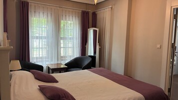 Habitación de hotel con cama, dos sillas, una mesita, espejo y ventana con cortinas.