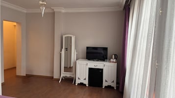 Habitación de hotel con piso de madera, un mueble blanco con televisor, un refrigerador pequeño y una puerta que da a otra habitación.