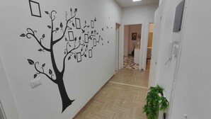 Un couloir orné d’une fresque murale représentant un arbre avec des feuilles et des boîtes, un plancher en bois, une porte avec une plante et un luminaire.