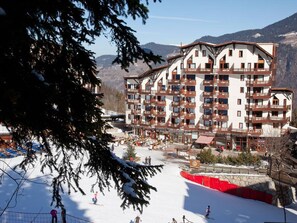 Um hotel com vários andares e varandas, encostado em pistas de esqui e uma paisagem montanhosa.