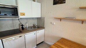 Cocina compacta con microondas, lavavajillas y pileta. Hay una mesa de madera y un estante pequeño sobre la pileta.