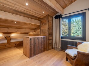 A wooden cabin interior with a loft bed, a wooden cabinet, a chair, and a window with a view of trees.