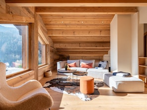 A cozy cabin interior with a wooden ceiling, a sofa set, a round coffee table, and a large window offering a mountain view.