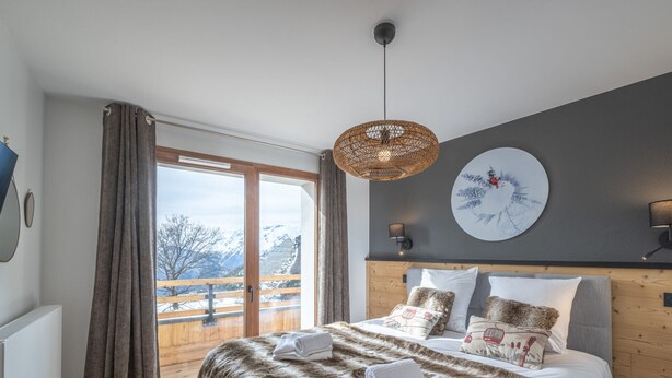 Une chambre moderne avec un grand lit, une tête de lit en bois, une vue sur les montagnes par une porte coulissante et un élément décoratif rond sur le mur.