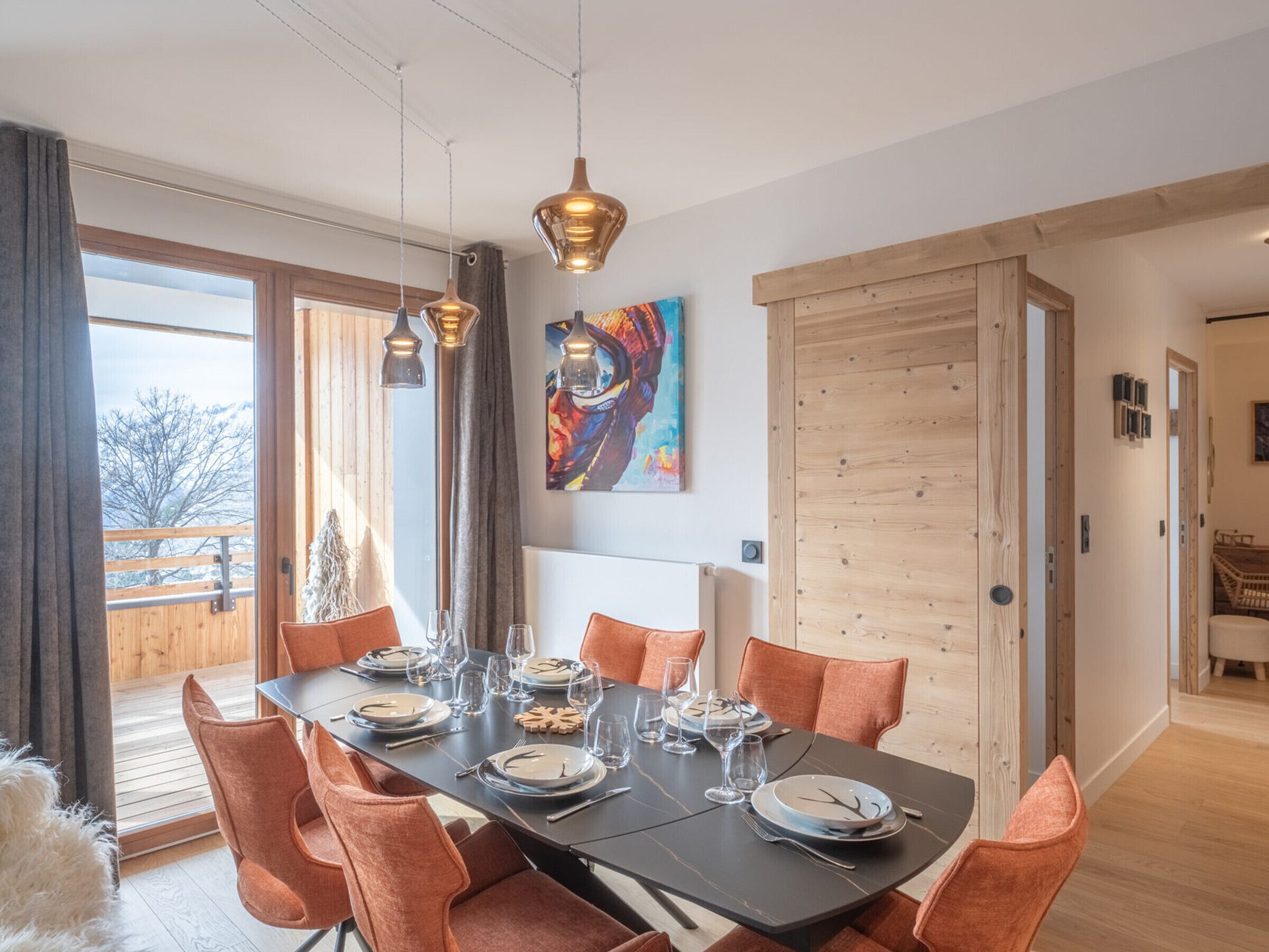 A dining area with a large table set for six, wooden flooring, and a view through the glass door.