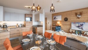 Une cuisine moderne avec des meubles en bois, un coin repas avec une table noire dressée pour six personnes, et un coin salon confortable agrémenté d’un plaid en fourrure.