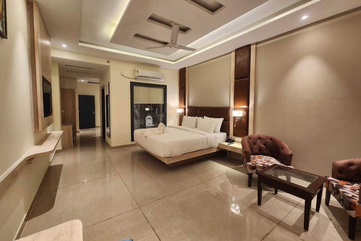 A modern hotel room with a large bed, a sitting area with a chair and table, a ceiling fan, and a bathroom visible through an open door.