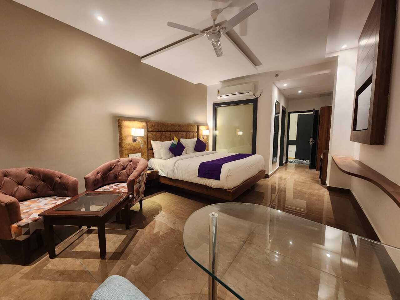Una habitación de hotel con cama, dos sillones, una mesa de centro de vidrio y un ventilador de techo.