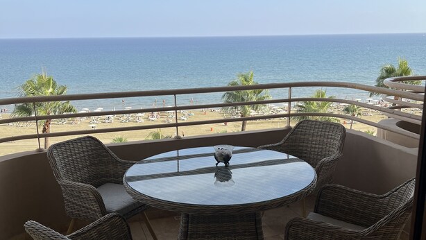 A balcony with wicker chairs and a glass table, overlooking a beach with palm trees and the ocean. - Finikoudes Beech front views Carisa Ithaki  (Larnaca)