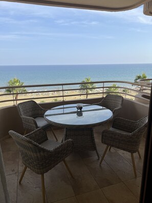 Un balcon agrémenté de fauteuils en osier et d’une table en verre, offrant une vue sur une plage bordée de palmiers et sur l’océan.