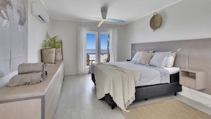Un dormitorio moderno con cama, mesitas de noche, ventilador de techo y vista al mar.