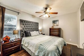 A bedroom with a tufted headboard, a bed with a striped bedspread, a nightstand with a lamp, a dresser, and a ceiling fan. - I36 - Apt|Yard|11mi to Fair Park|Sleeps 4 (Irving)