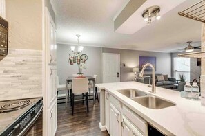 A modern kitchen with a central island, a dining area with a chandelier, and a living room with a sofa and a TV. - I36 - Apt|Yard|11mi to Fair Park|Sleeps 4 (Irving)