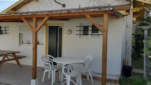 Un espace extérieur abrité avec une table de pique-nique et des chaises, un bâtiment blanc doté de fenêtres et d’une porte, et une voiture garée à proximité.