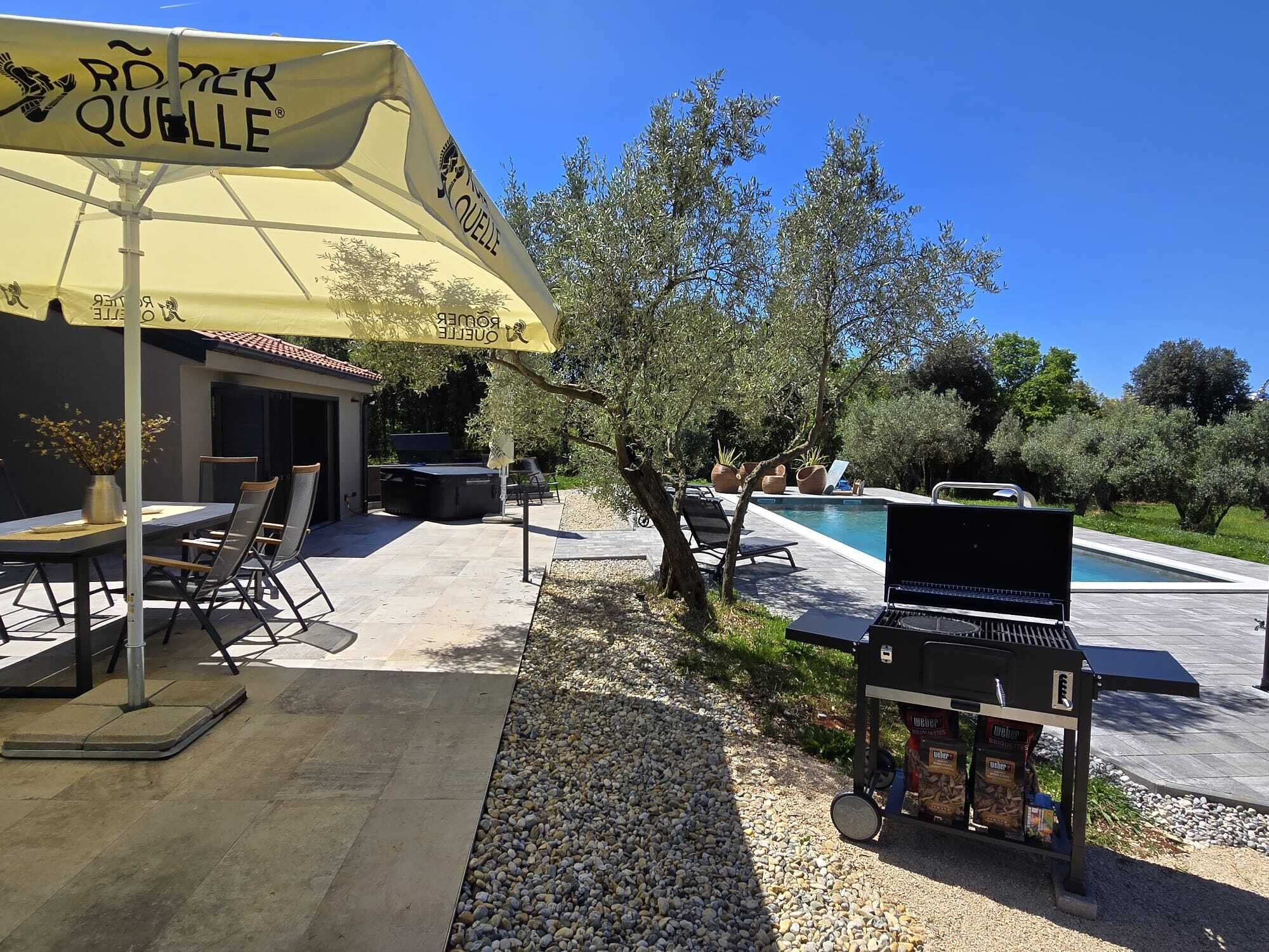 An outdoor patio area with a pool, a grill, and a table with chairs under a yellow umbrella.