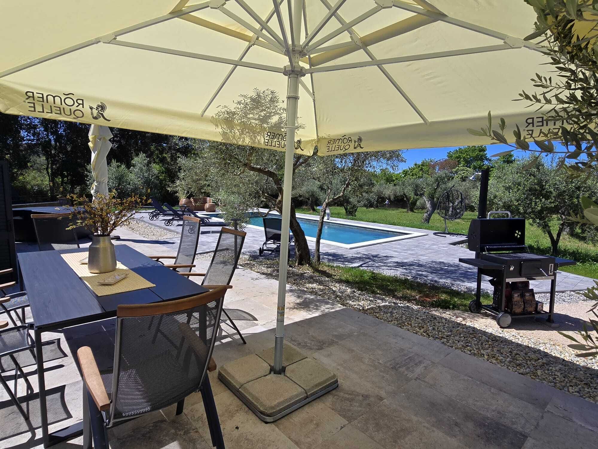 An outdoor patio with a table and chairs under a large umbrella, a pool, and a barbecue grill.