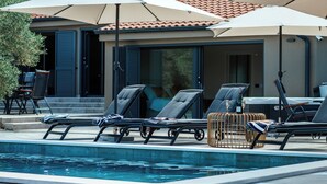 A poolside area with lounge chairs and umbrellas, a table, and a view of a building with a tiled roof. - Olive Garden Paradise with Swimming pool (Rovinj)