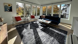 Ein modernes Wohnzimmer mit Sofa, Sessel, Couchtisch und einem großen Fenster mit Blick auf Bäume und ein Haus.