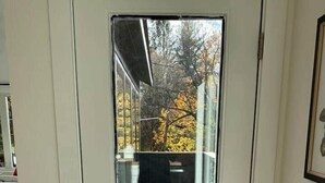 Eine weiße Tür mit Glasfenster, die zu einem überdachten Außenbereich mit Blick auf Bäume und ein Gebäude im Hintergrund führt.