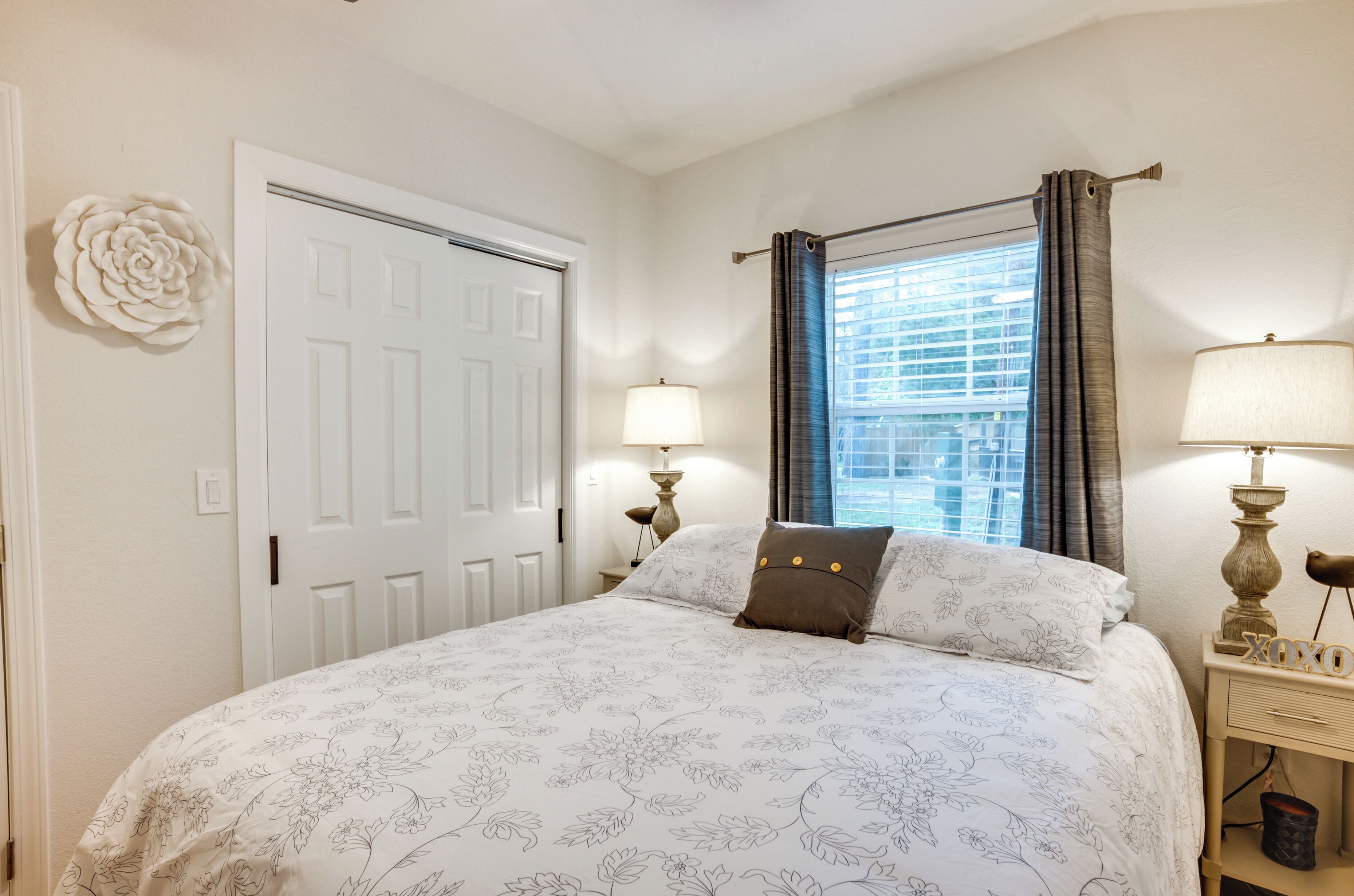 A bedroom with a bed, two bedside lamps, a window with curtains, a door, and a wall decoration.