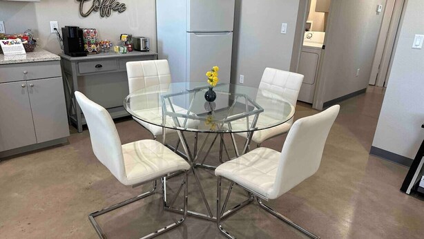 A modern kitchen with a glass dining table, four white chairs, a refrigerator, and a coffee sign on the wall.