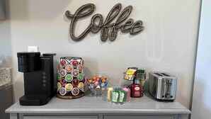 A coffee station with a coffee maker, a coffee-themed wall sign, a display of coffee pods, a toaster, and a coffee grinder.