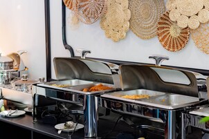 A buffet setup with multiple stainless steel chafing dishes containing various foods, a coffee machine, and decorative wall hangings.