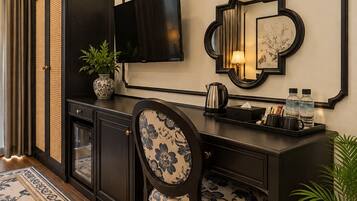 A modern hotel room with a dark wood desk, a flat-screen TV, a decorative mirror, and a floral-patterned chair.