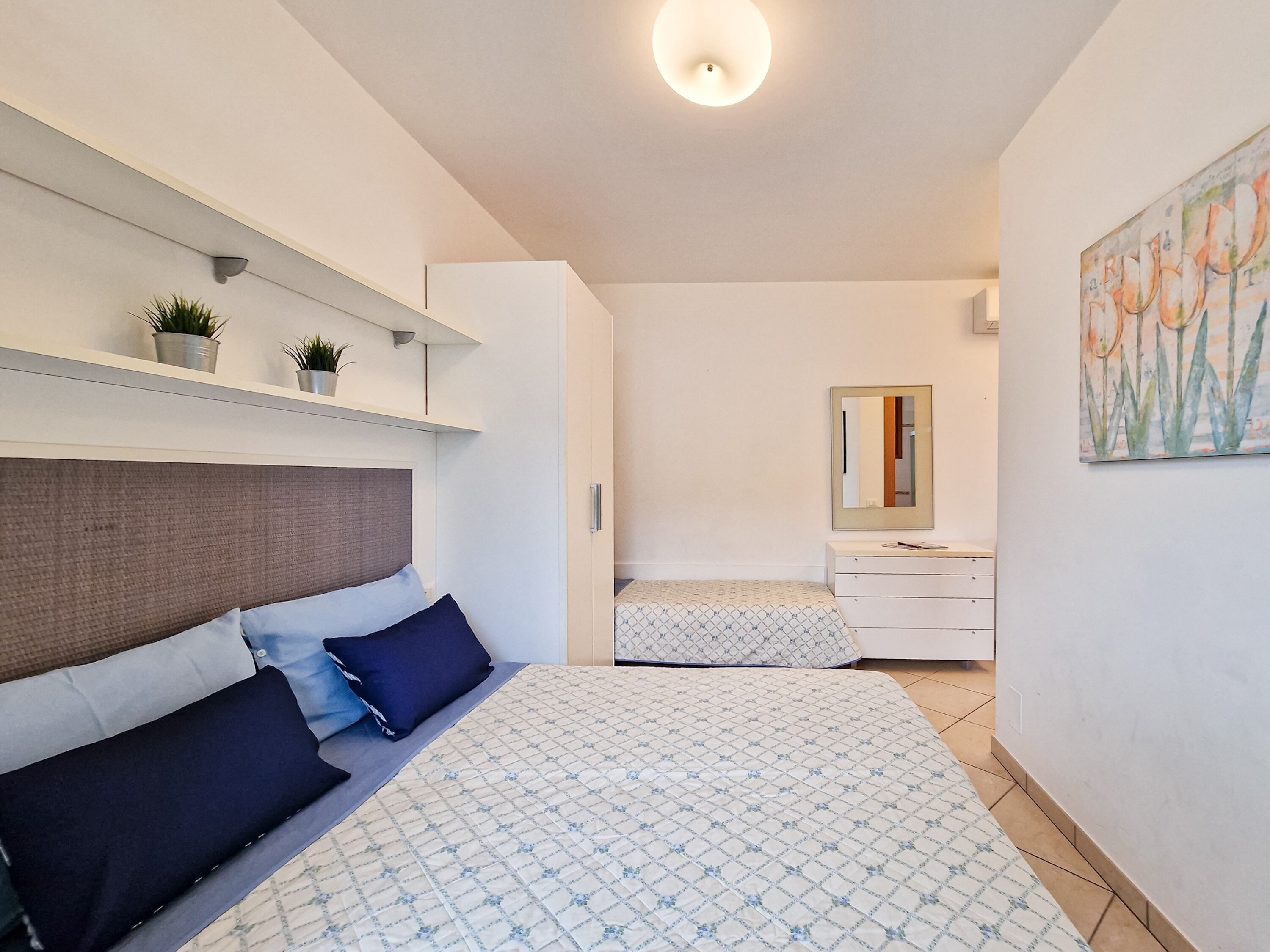 A bedroom with two beds, a bedside table, a dresser, a mirror, and wall-mounted shelves with plants.